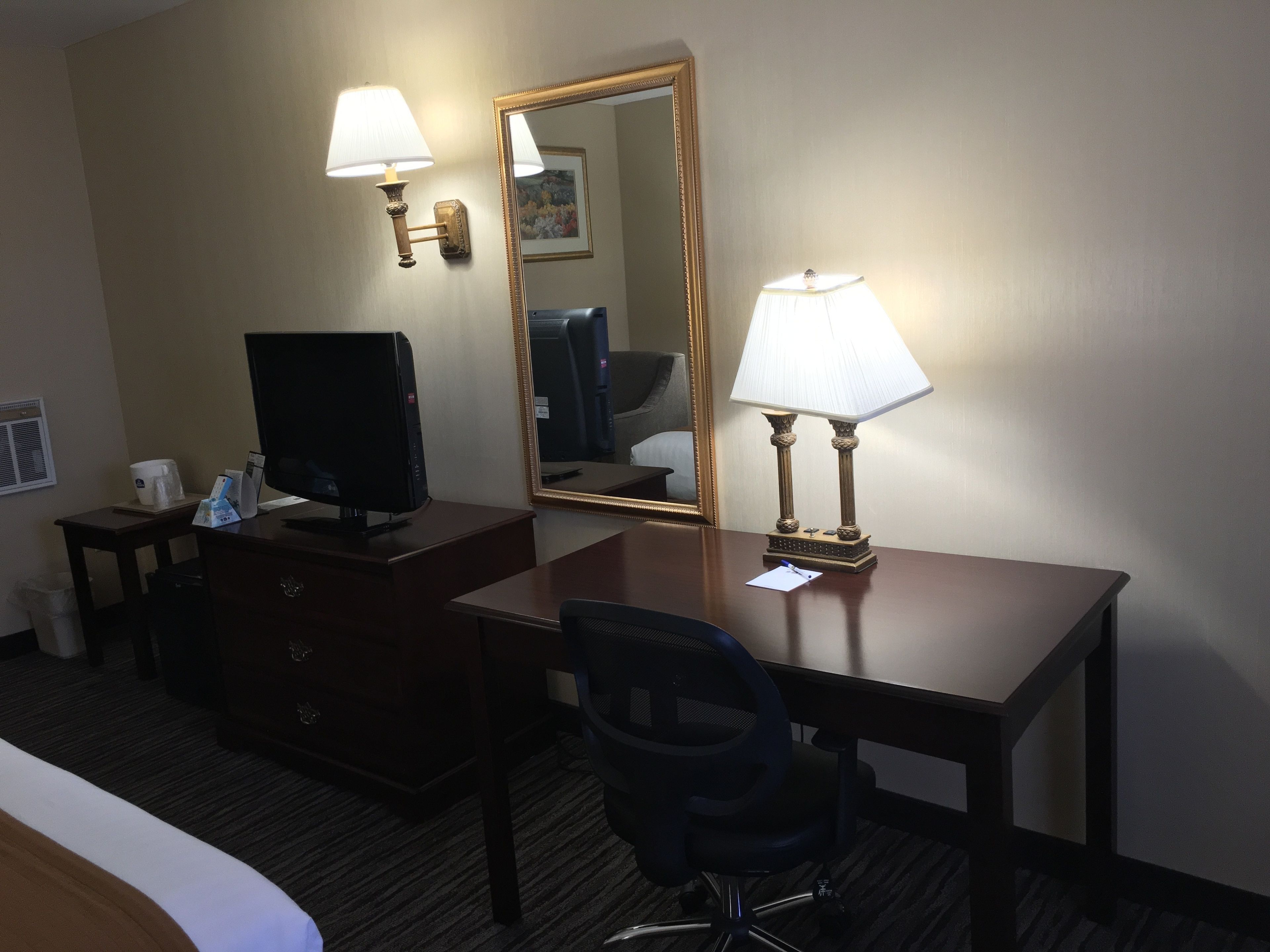 Habitación de hotel con escritorio, dos lámparas, televisor, espejo y una silla.