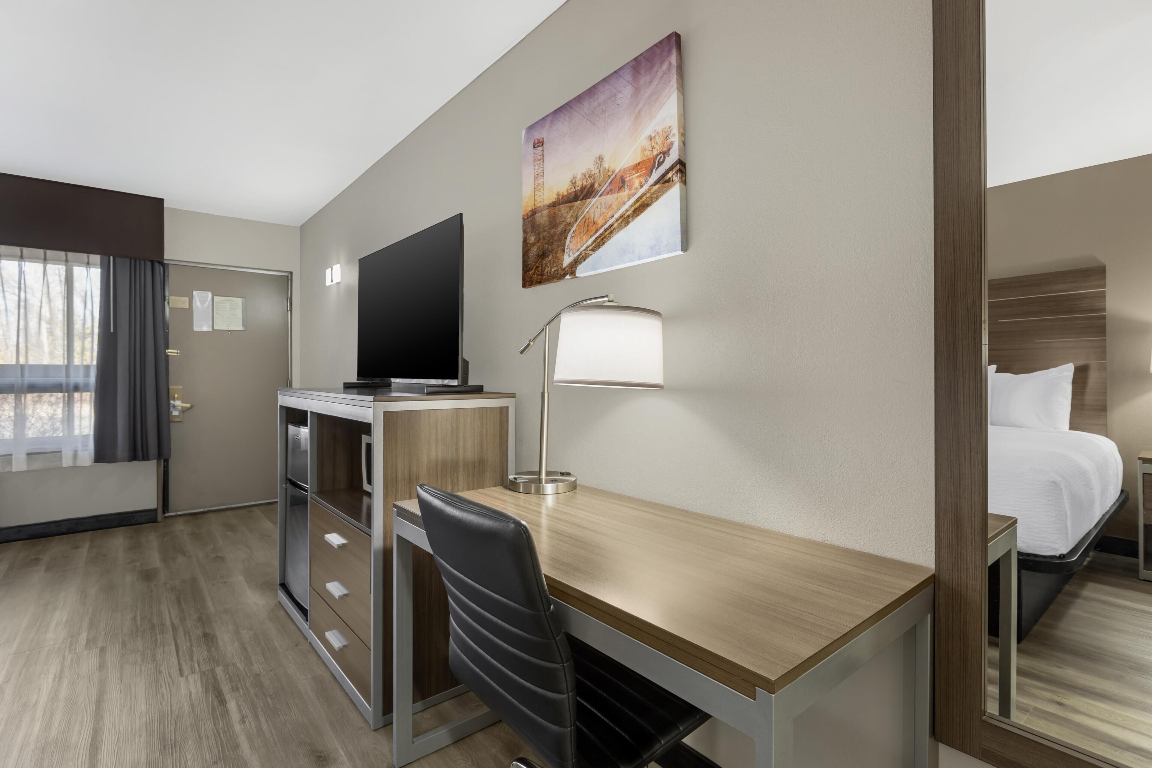 A hotel room with a desk, chair, bed, TV, lamp, and a painting on the wall.