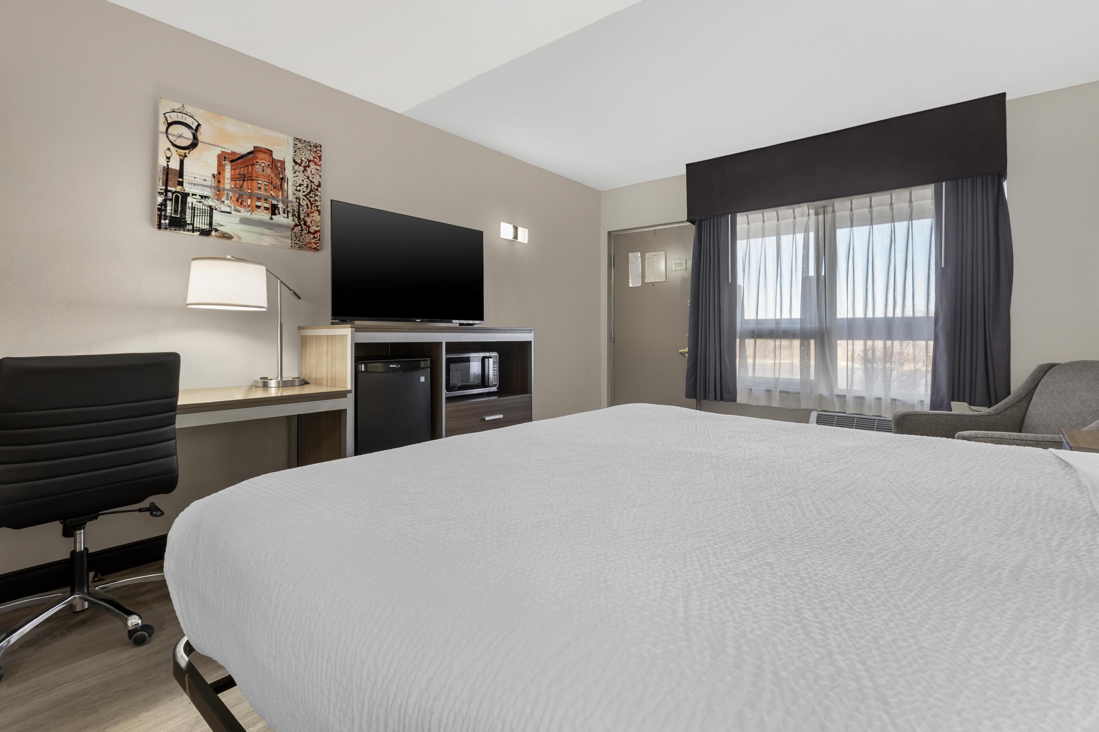 Una habitación de hotel con cama, escritorio, silla, televisión y ventana con cortinas.