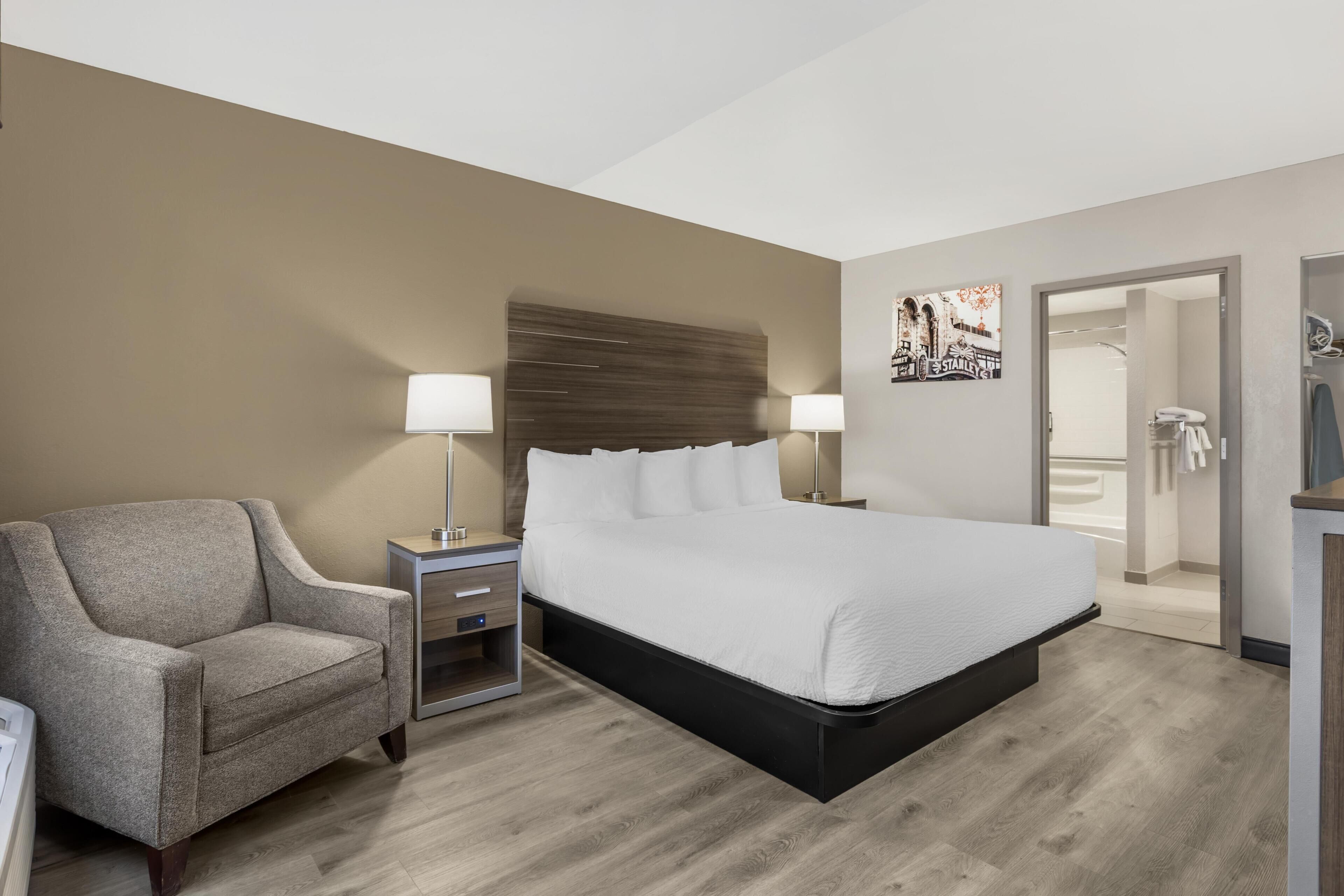 A hotel room with a bed, a chair, a nightstand, a lamp, and a painting on the wall.