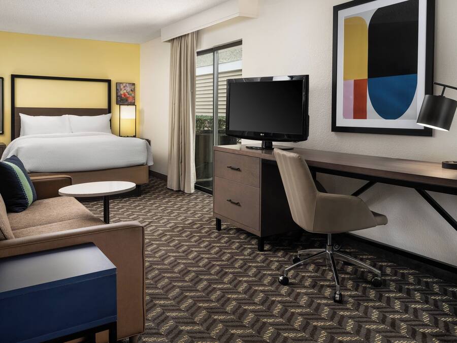 A hotel room with a bed, a desk, a chair, a TV, a lamp, and a painting on the wall.