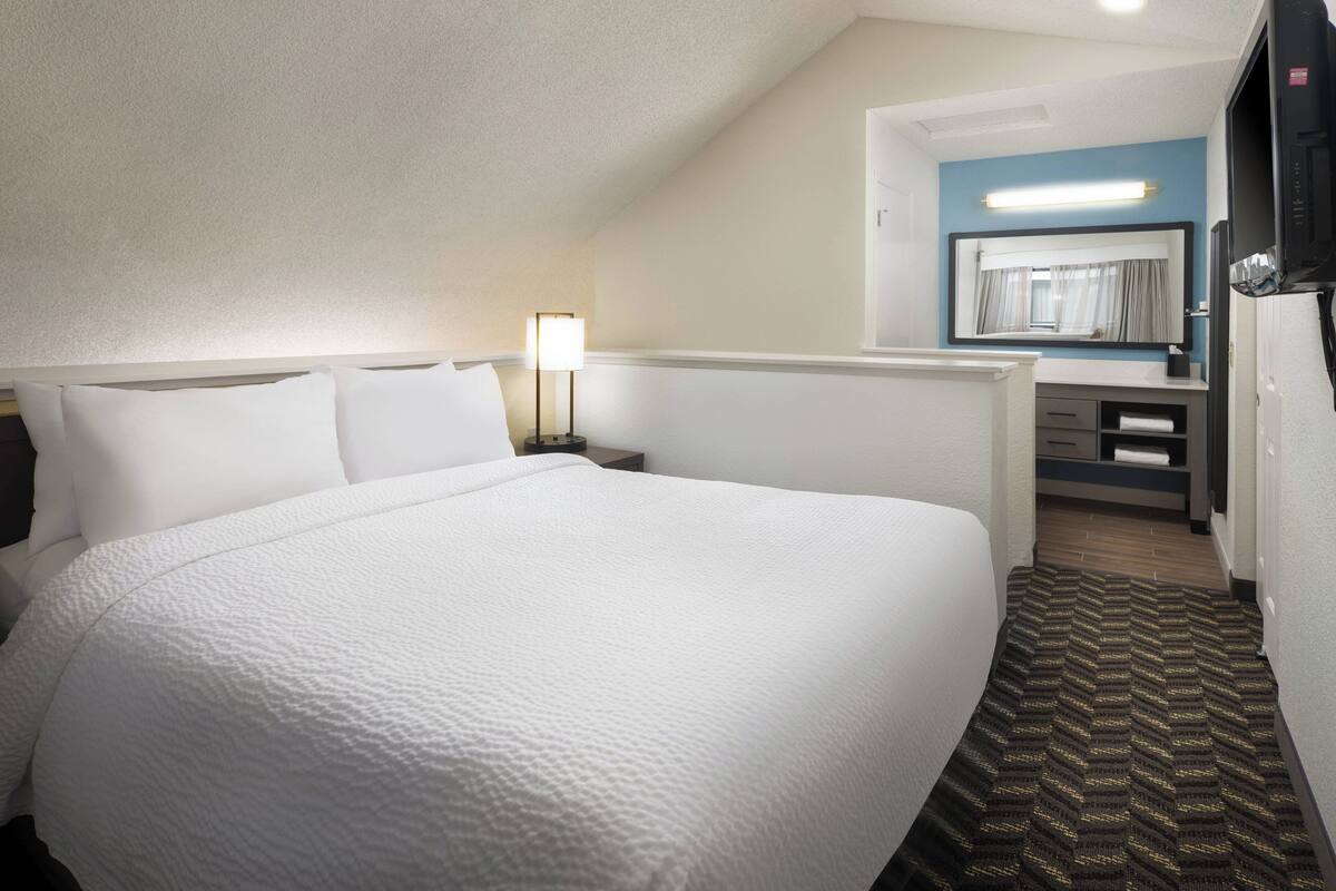 Una habitación de hotel con una cama grande, una mesita de noche con lámpara, un espejo, un televisor y un escritorio pequeño con cajones.