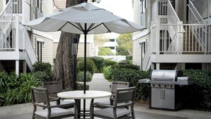 Een terras buiten met een ronde tafel, vier stoelen, een grote parasol en een barbecue.