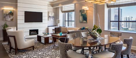 A modern hotel room with a large window, a flat-screen TV mounted on the wall, a round dining table with chairs, and a patterned rug.