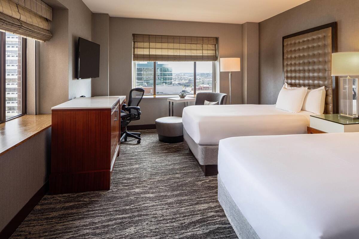 A hotel room with two beds, a desk, a chair, a small table, and a TV.