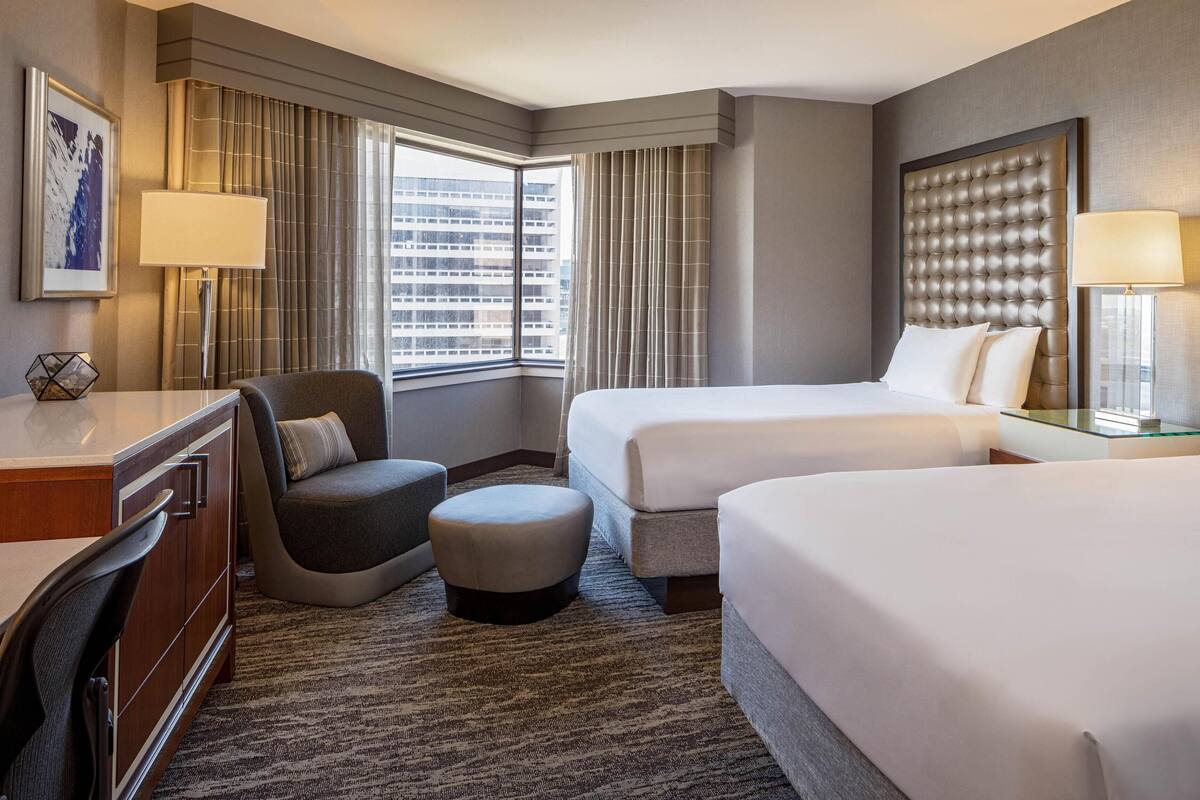 A hotel room with two beds, a desk, a chair, and a large window with curtains.