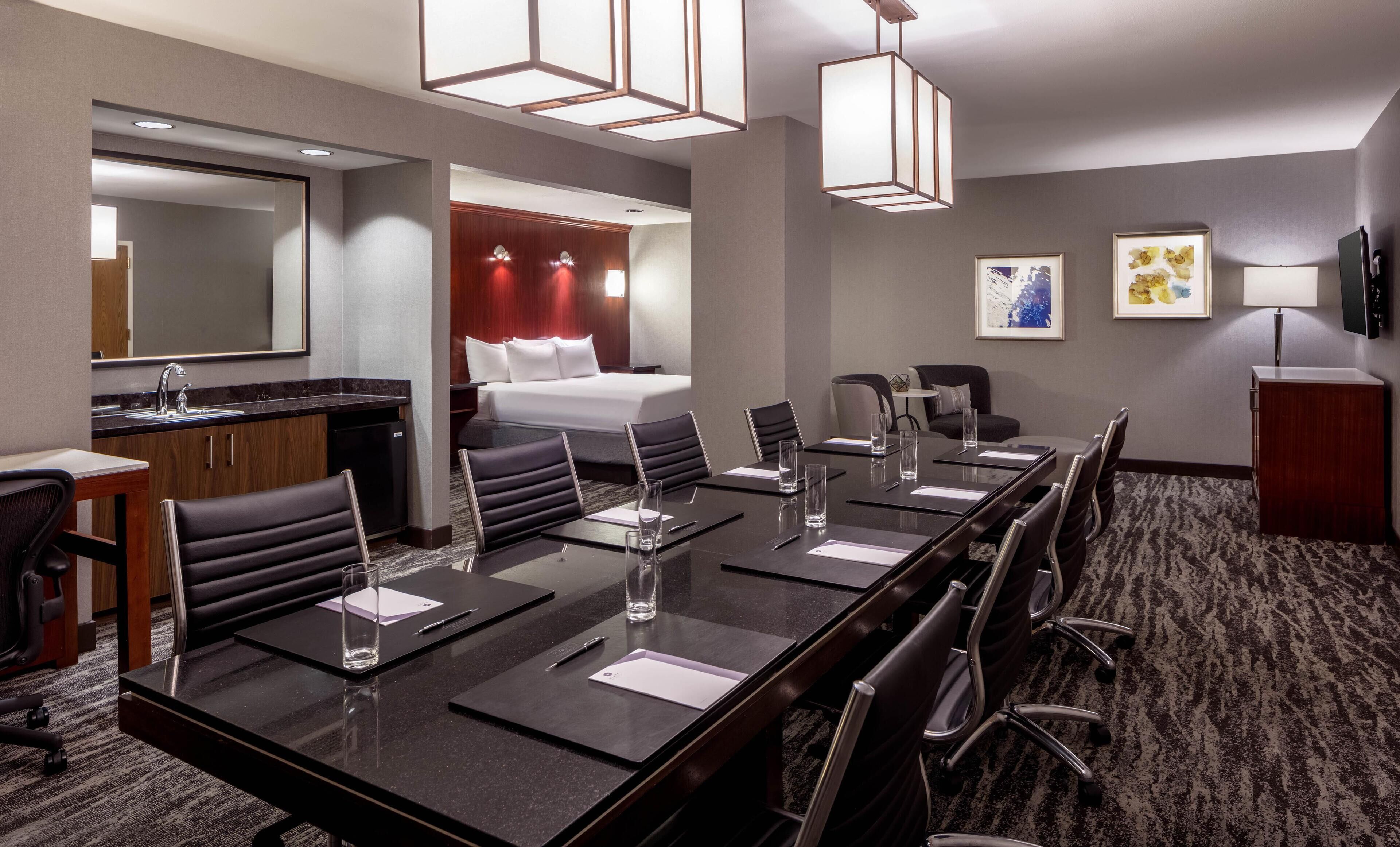 A hotel conference room with a long table, chairs, a bed, and a bathroom.