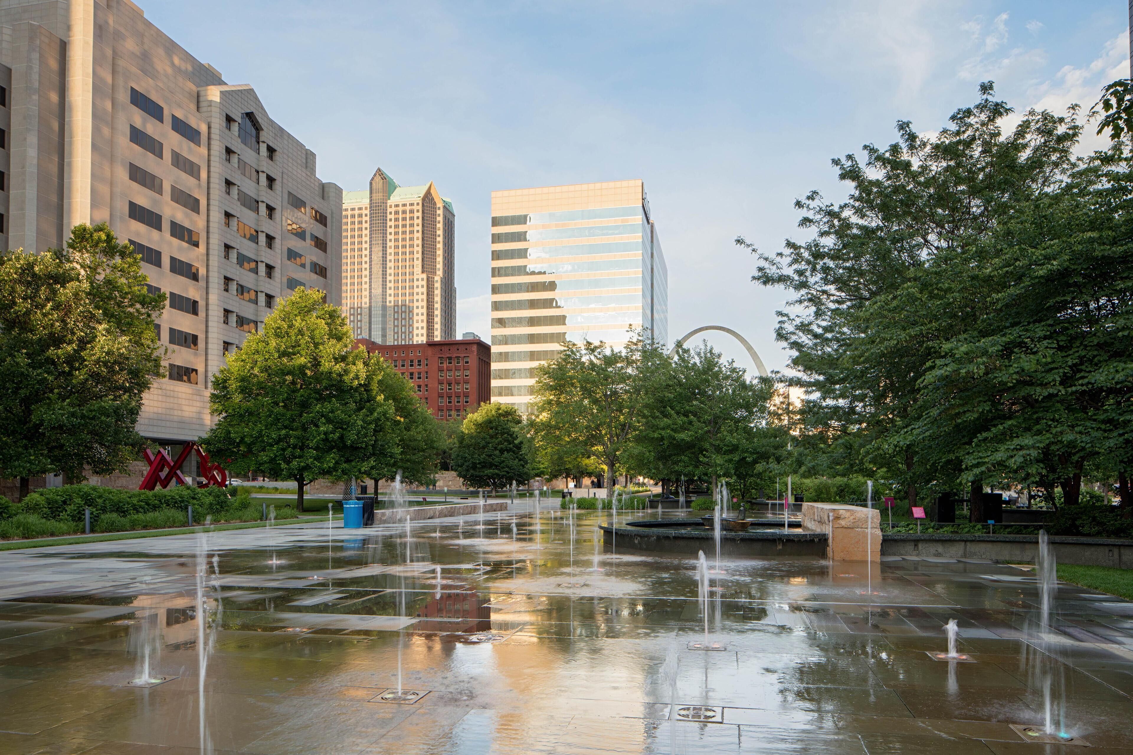 A public square with a fountain, surrounded by trees and modern buildings.