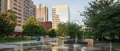 A public square with a fountain, surrounded by trees and modern buildings.