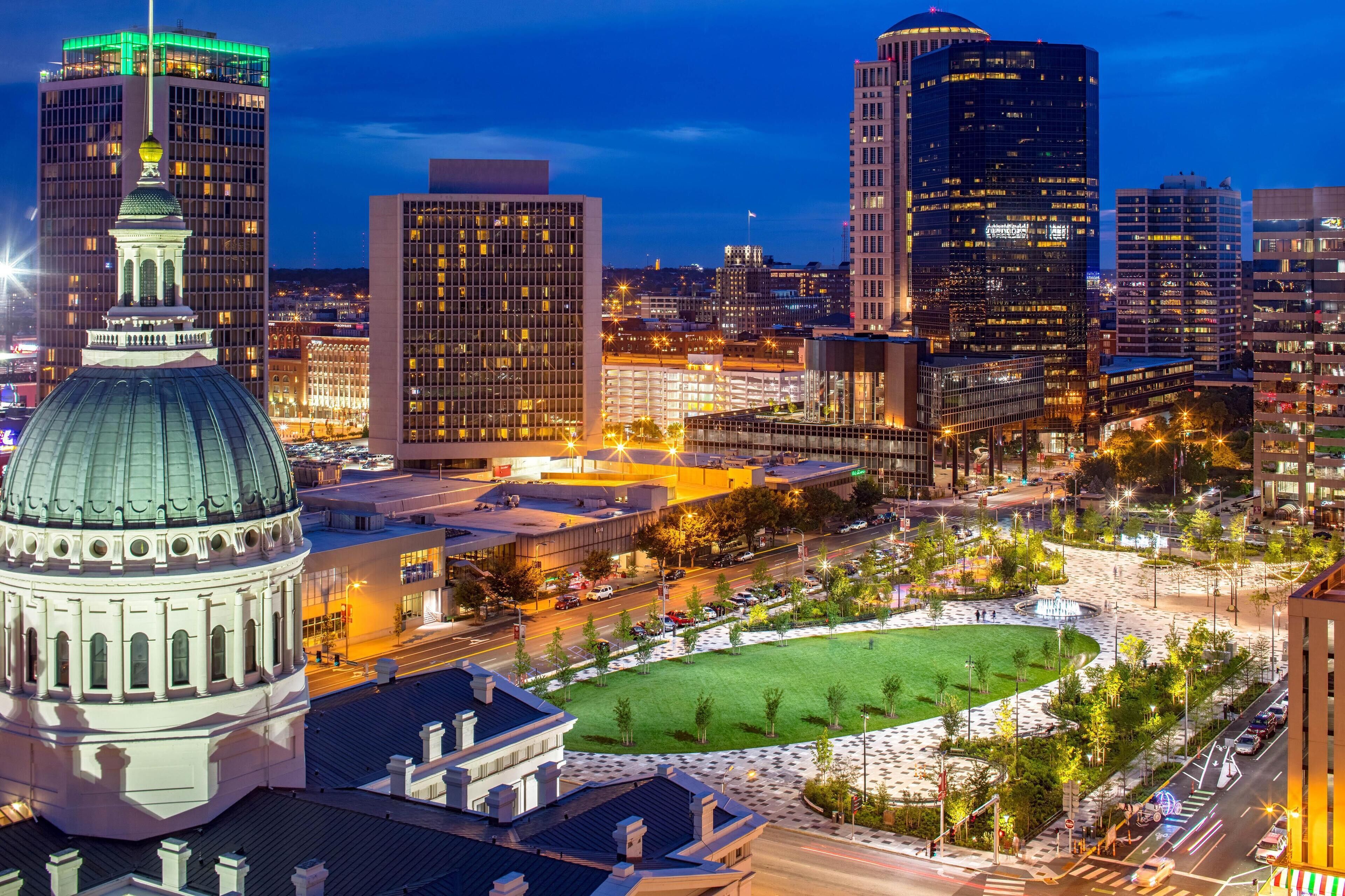 A cityscape with modern buildings, a prominent dome structure, and a well-lit public square.