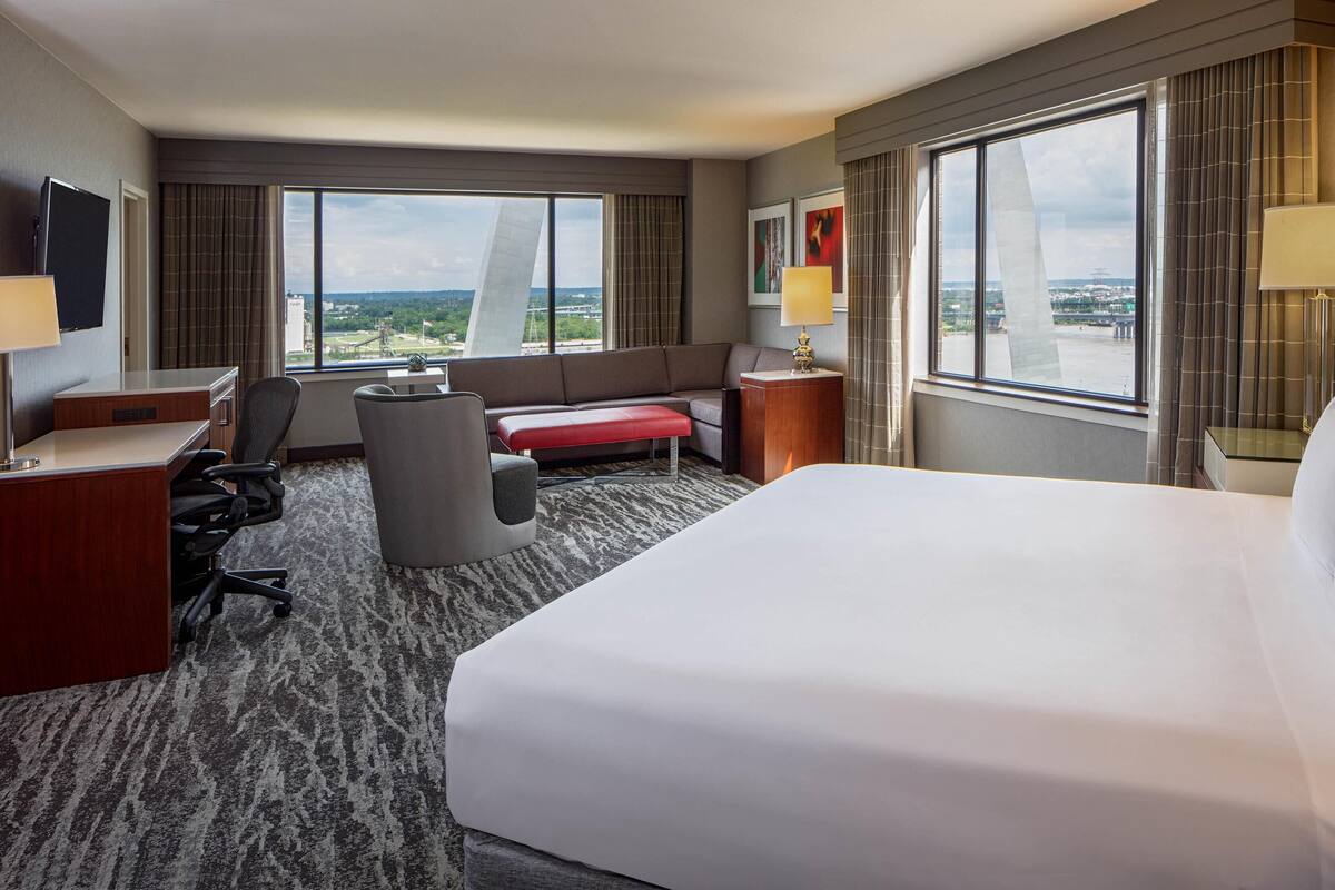 A hotel room with a large bed, a desk, a chair, a sofa, and a view of the cityscape through large windows.