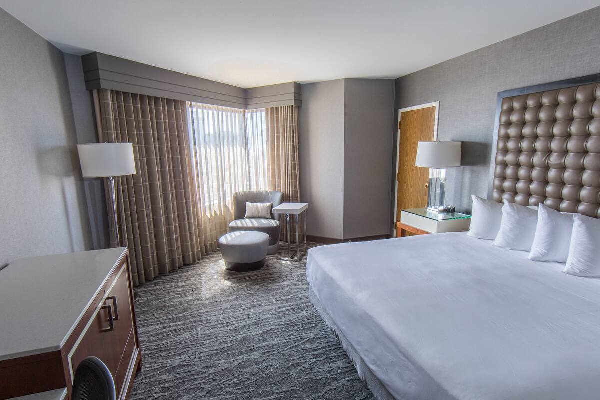 A modern hotel room with a large bed, a seating area with a chair and ottoman, a small table, and a large mirror.