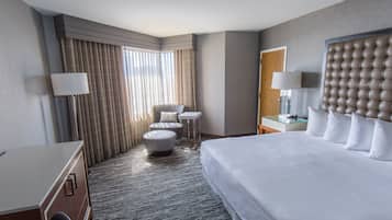 Moderni hotellihuone, jossa on suuri sänky, oleskelualue tuolilla ja jalkarahilla, pieni pöytä ja suuri peili.