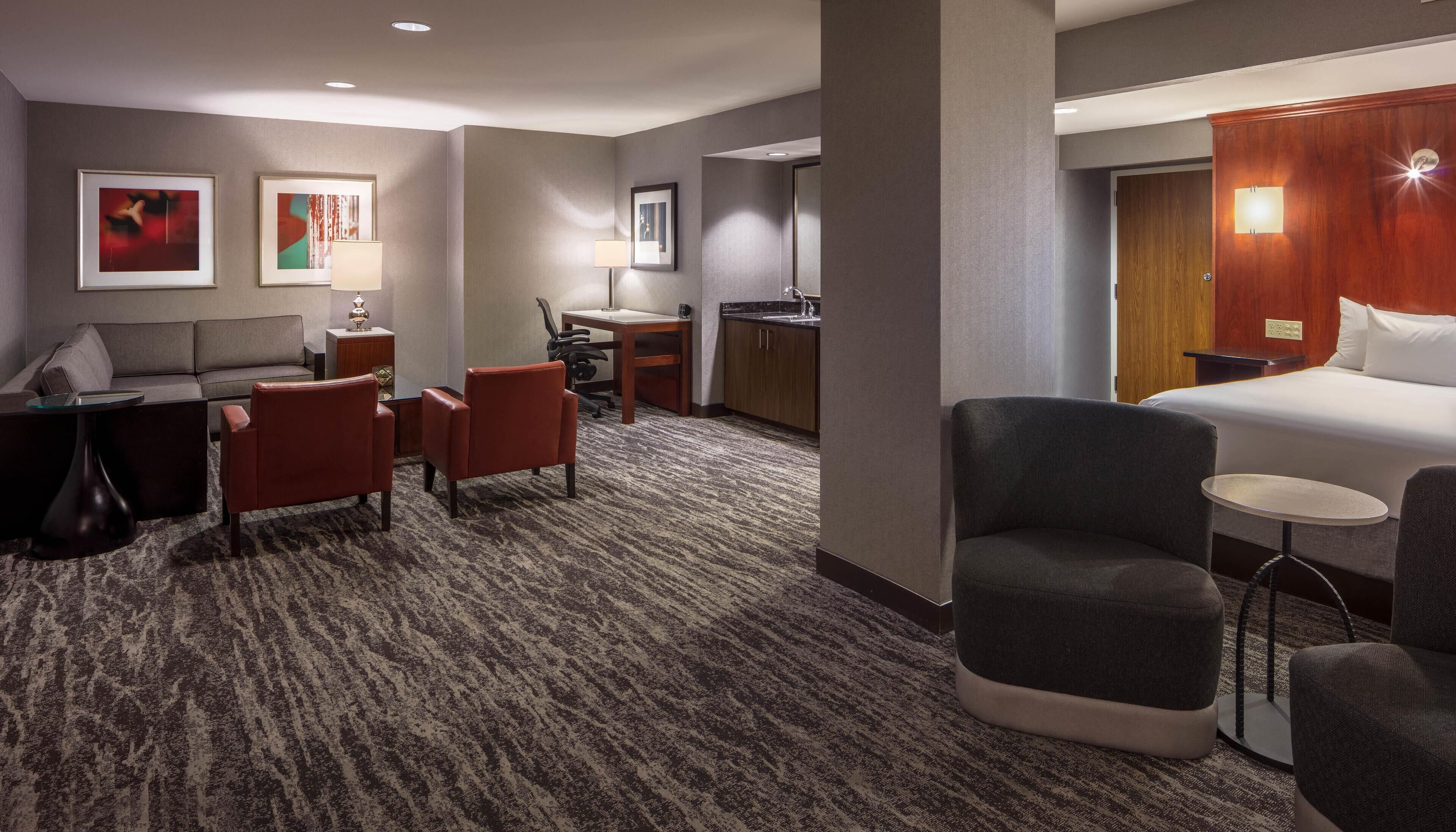 A hotel room with a bed, two armchairs, a small table, a desk with a chair, a sofa, and a carpeted floor.