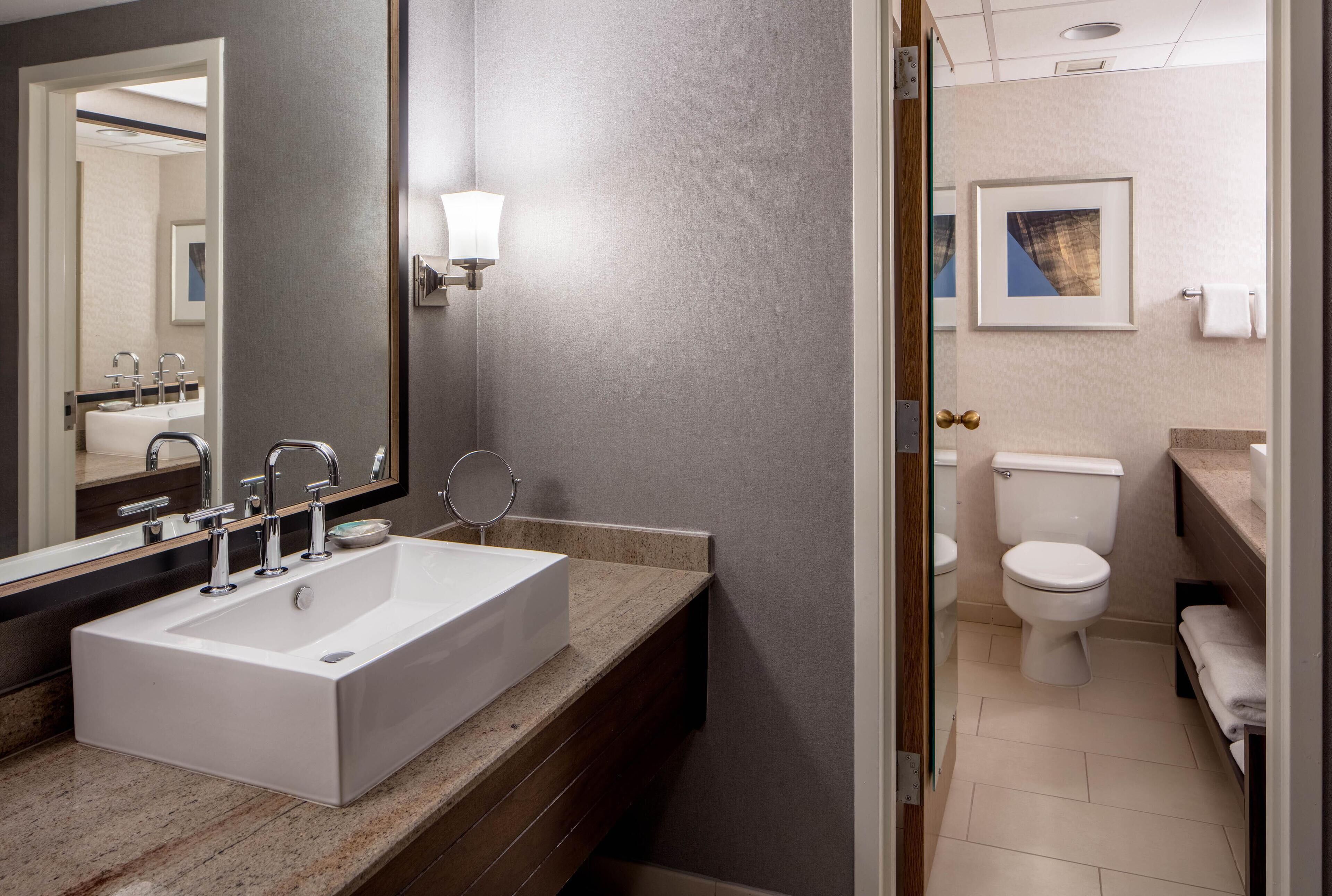 A modern bathroom with a rectangular sink, a mirror, a wall-mounted light, a toilet, and a framed picture on the wall.