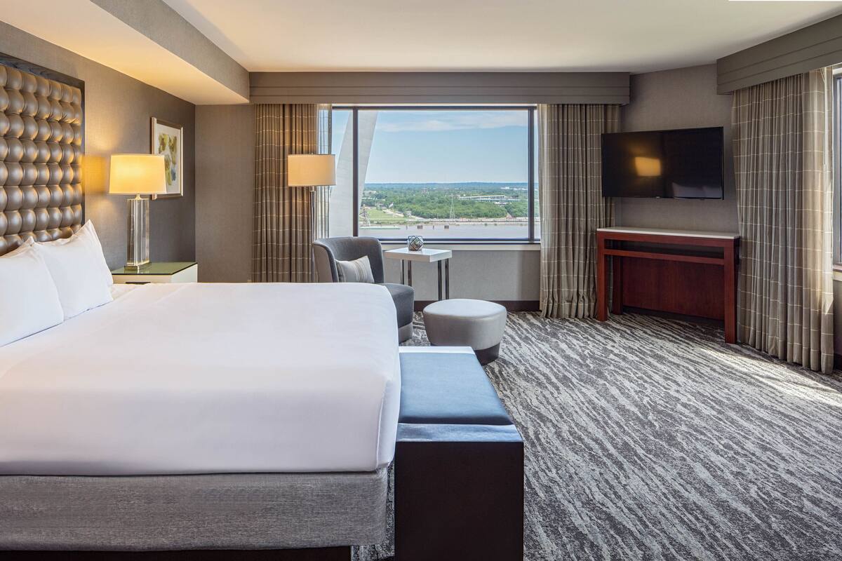 A hotel room with a large bed, a desk, a chair, a TV, and a view of a cityscape.