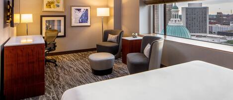 A hotel room with a bed, two armchairs, a desk, and a view of a cityscape through a large window.