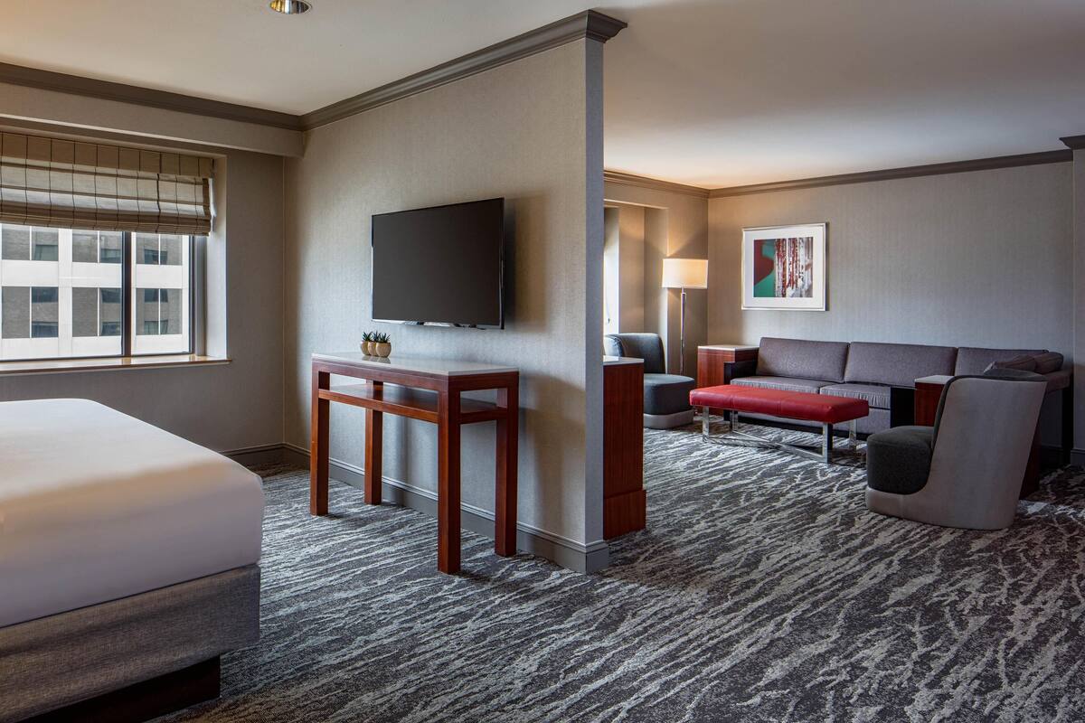 A hotel room with a bed, a TV, a sofa, a red bench, and a small table.