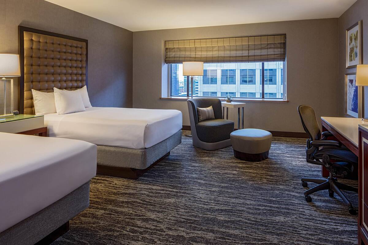 A hotel room with two beds, a desk, a chair, an ottoman, and a large window with a view of buildings.
