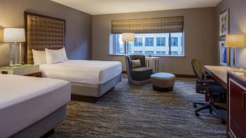 A hotel room with two beds, a desk, a chair, an ottoman, and a large window with a view of buildings.