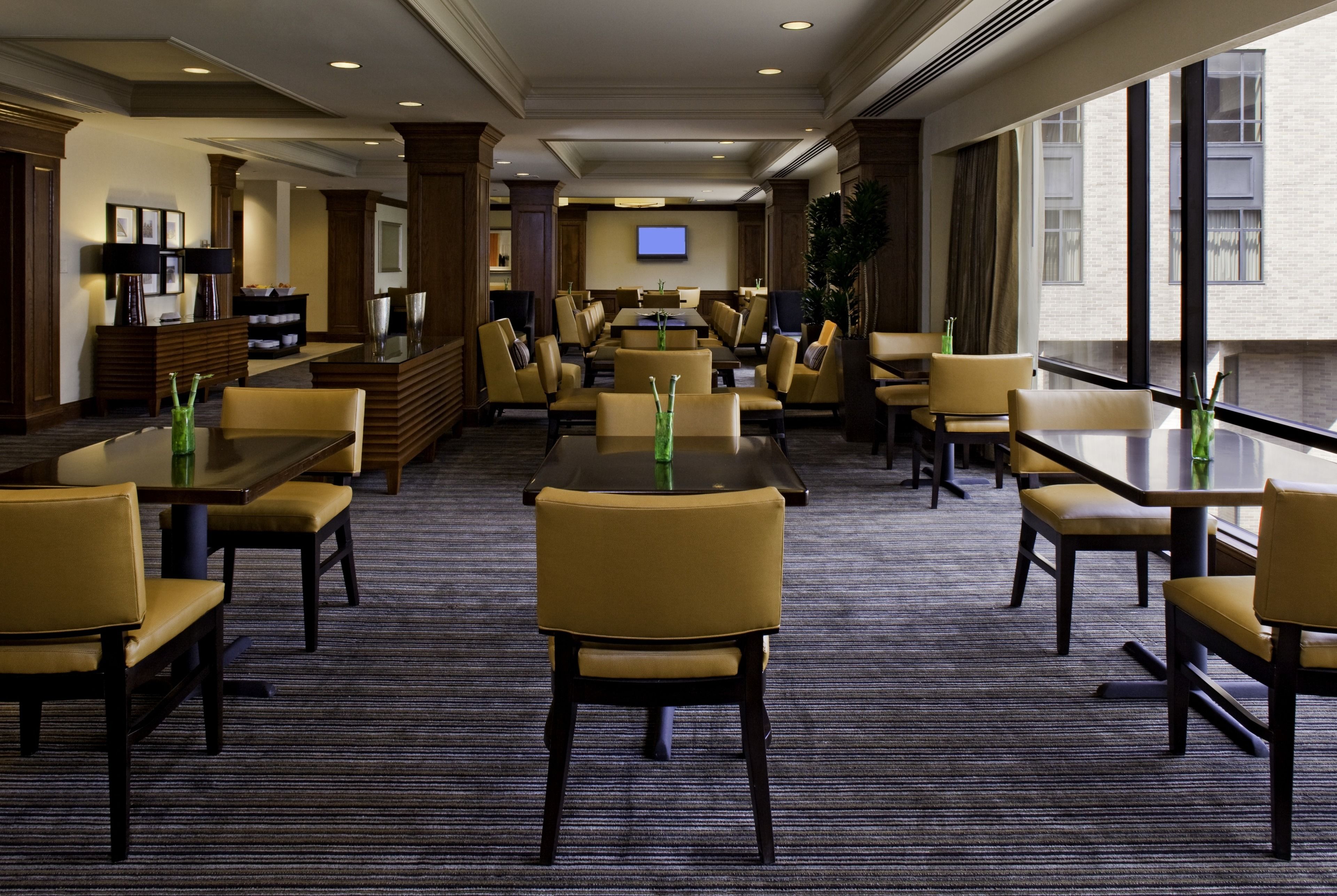 A hotel lobby with multiple seating areas, a television mounted on the wall, and large windows providing a view of the outside.