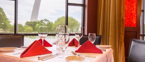 Mesa de comedor dispuesta con servilletas rojas, copas de vino y cubiertos, con vista a árboles a través de amplias ventanas.