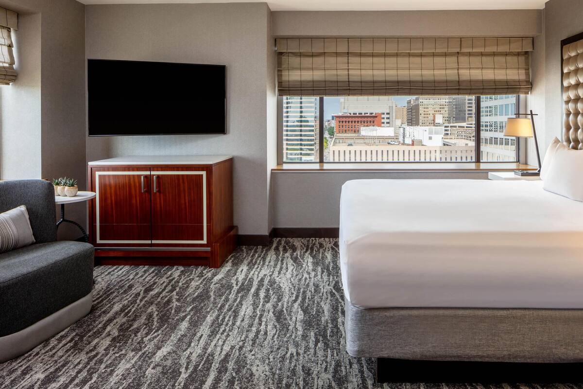 A hotel room with a large bed, a TV mounted on the wall, a small table with a lamp, and a view of the city through the window.