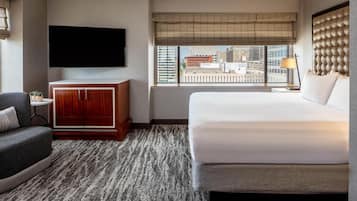 Habitación de hotel con una cama grande, un televisor montado en la pared, una mesita con lámpara y vistas a la ciudad desde la ventana.