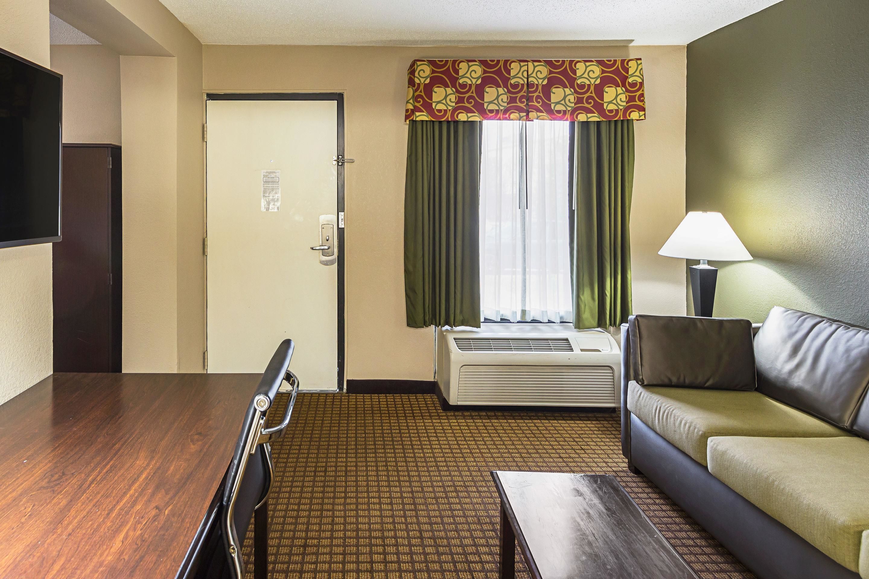 A hotel room with a wooden dining table, a sofa, a TV, a window with curtains, and an air conditioning unit.