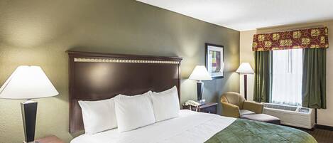 A hotel room with a large bed, two bedside lamps, a chair, a desk with a telephone, and a window with curtains.
