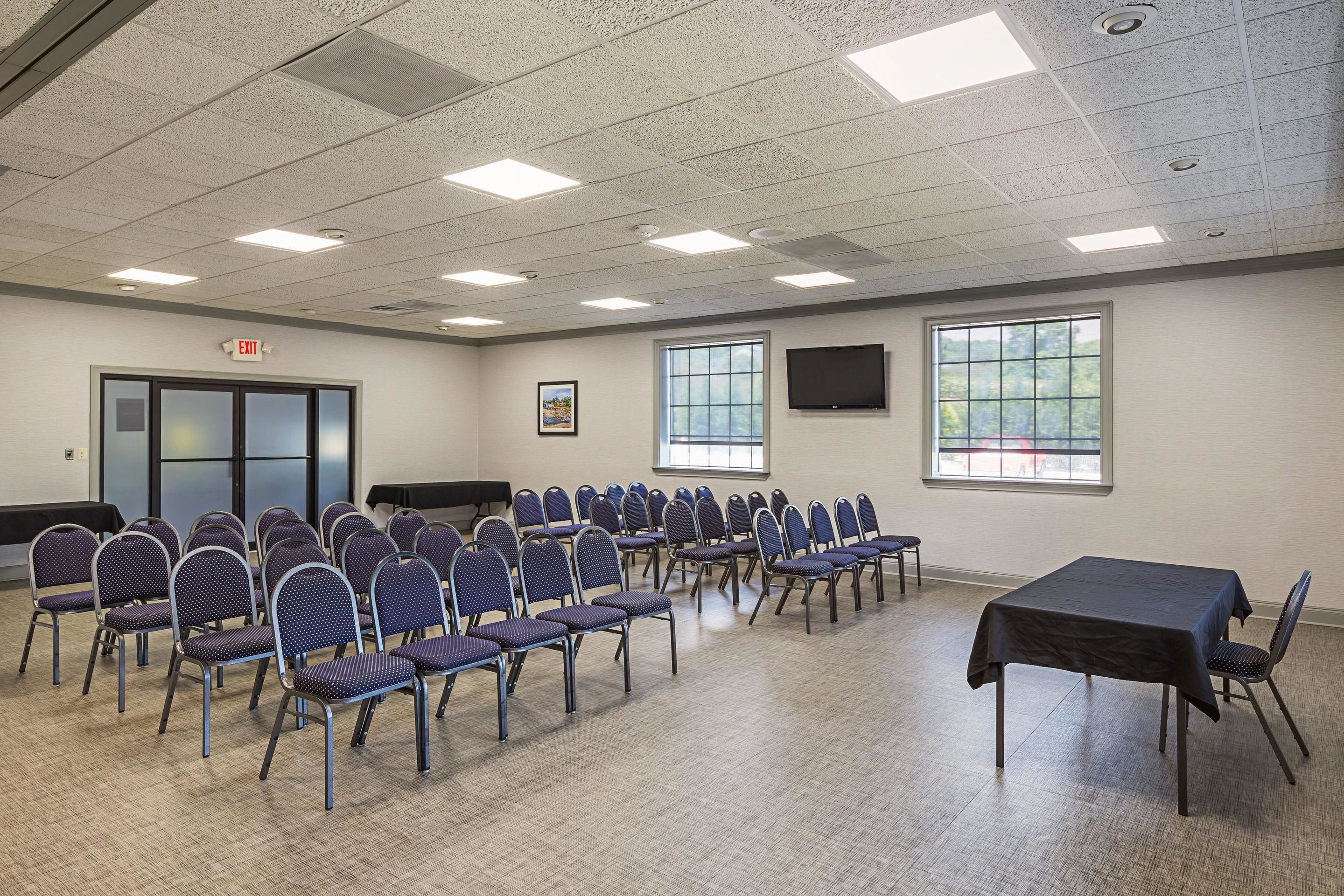 A conference room with rows of chairs, a table, a projector screen, and windows.
