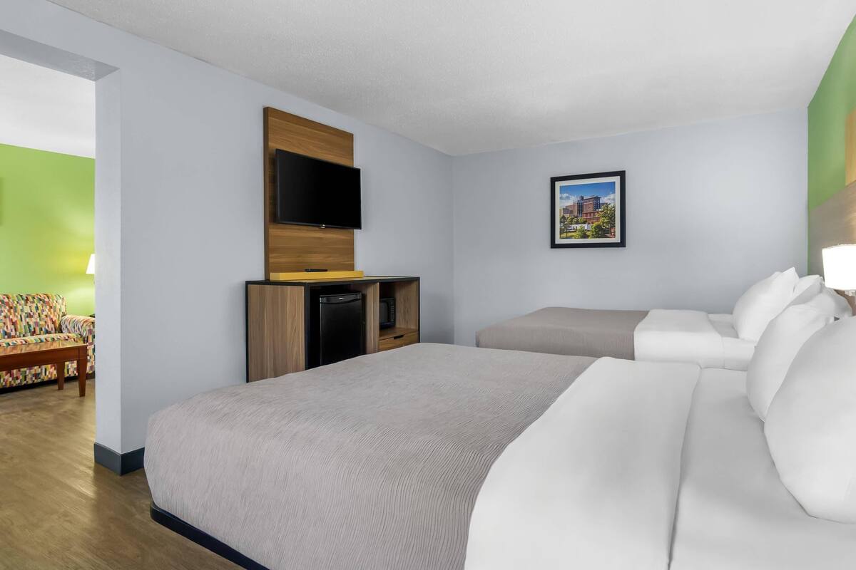 A hotel room with a large bed, a sofa, a flat-screen TV mounted on a wooden panel, a small desk with a computer, and a framed picture on the wall.