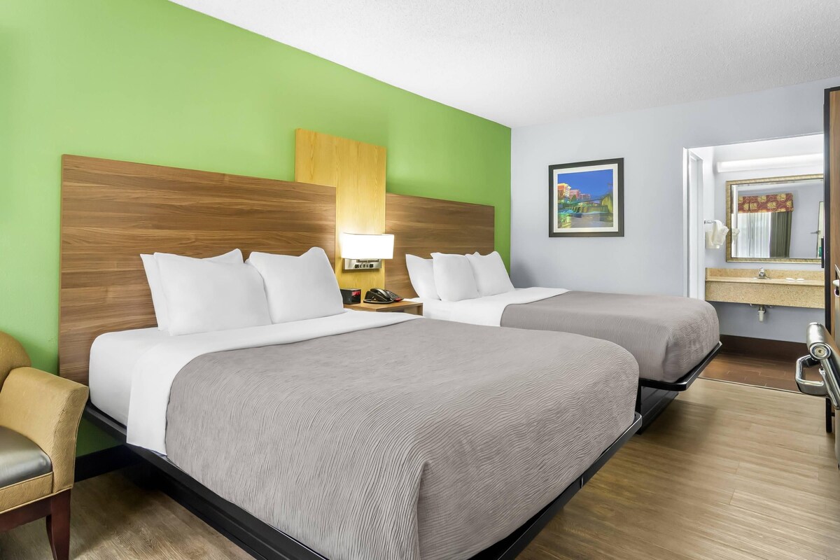 A hotel room with two beds, a wooden headboard, a green wall, a painting, and a bathroom with a mirror and sink.