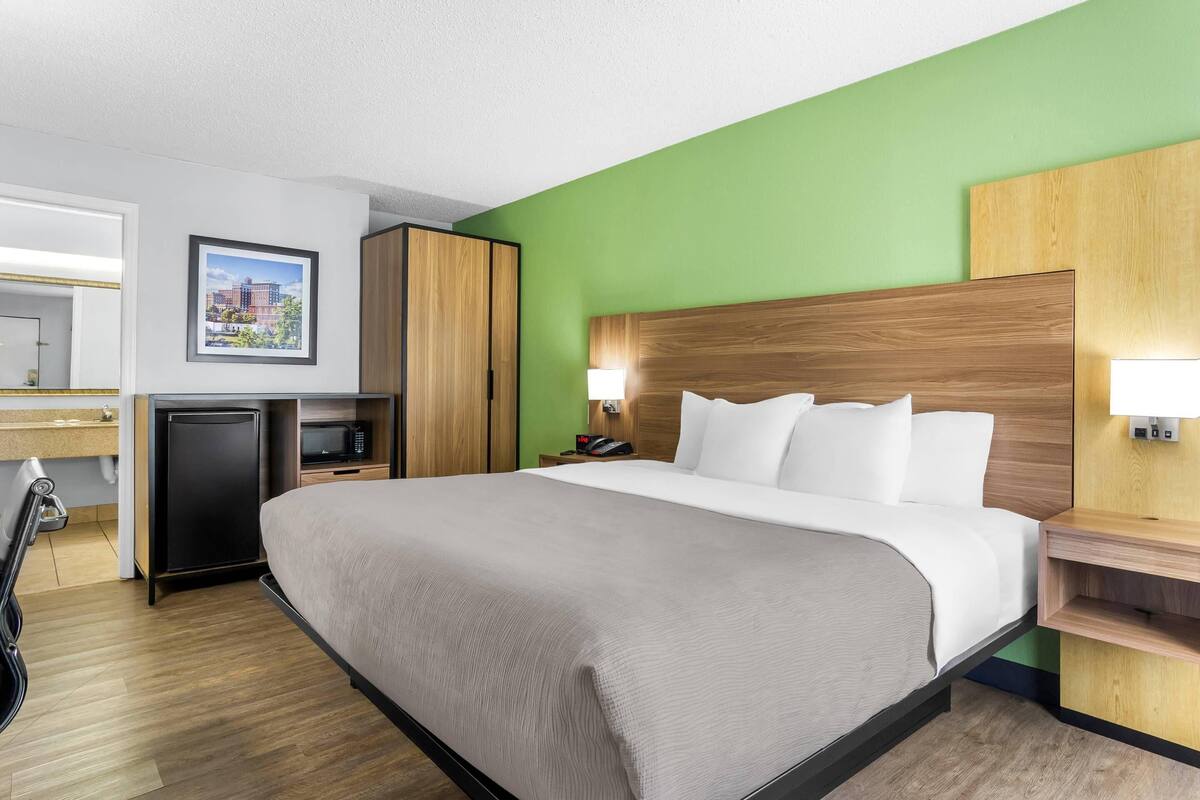 A hotel room with a large bed, a desk with a computer, a small fridge, and a green accent wall.
