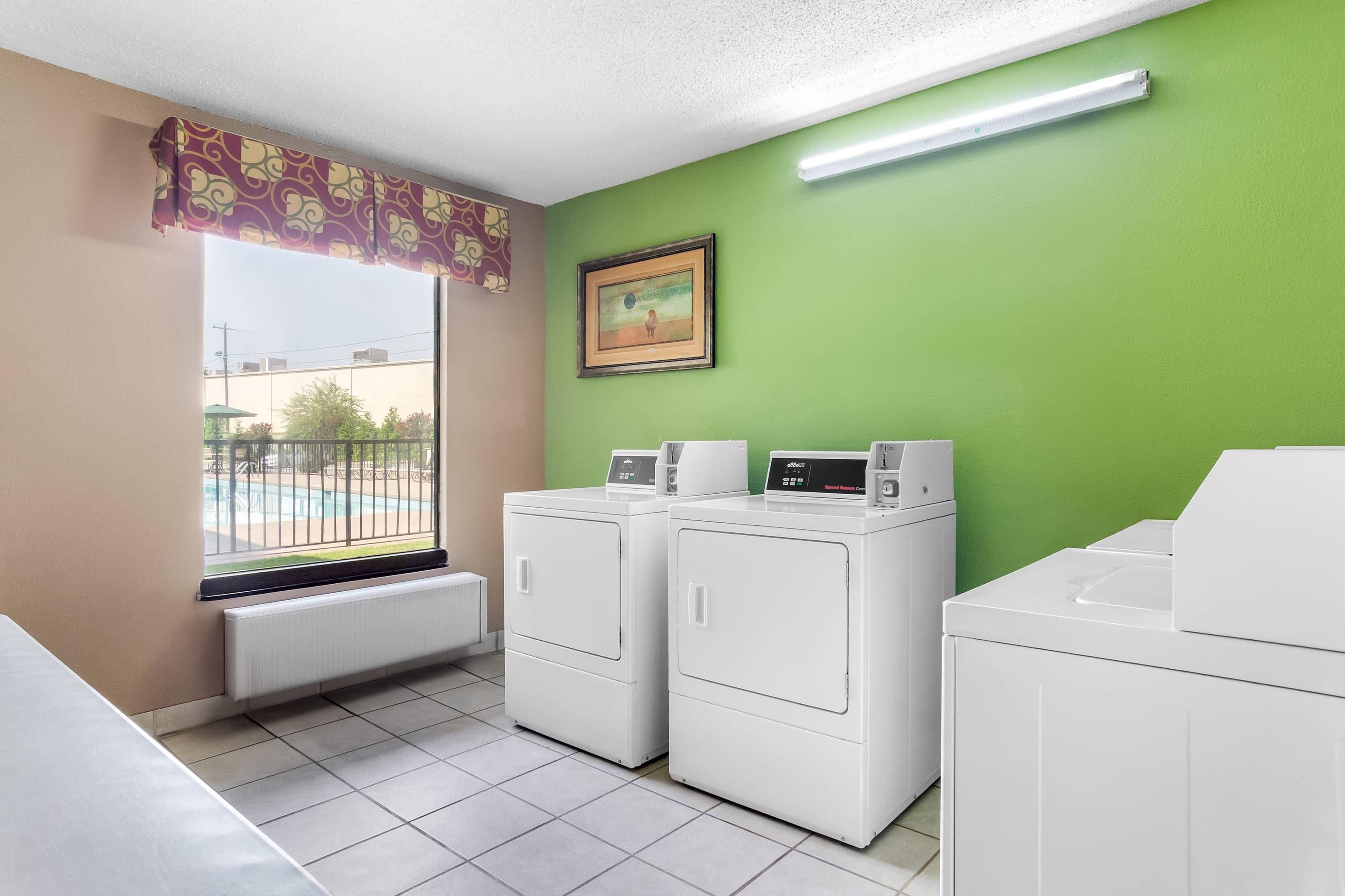Una sala de bugaderia amb dues rentadores blanques, un radiador, una finestra que donava a una piscina i una paret verda.