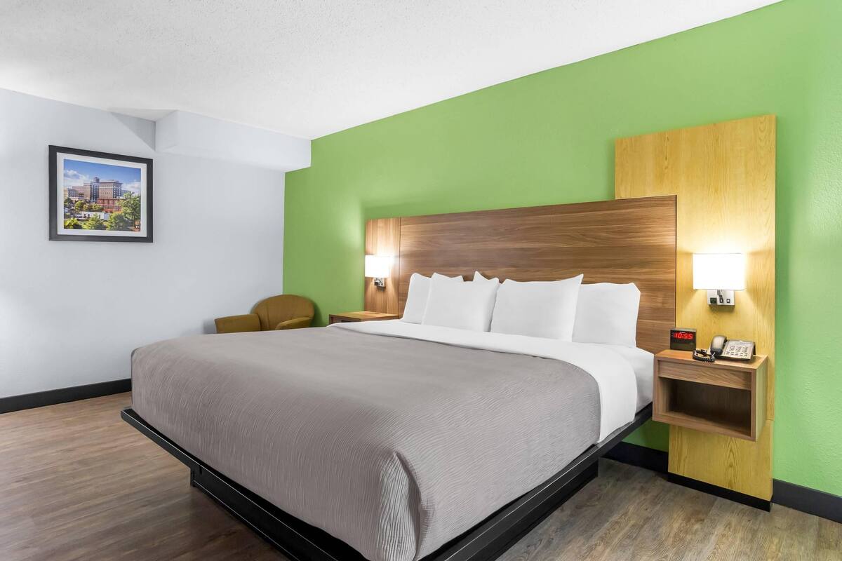A hotel room with a bed, a nightstand with a lamp, a chair, and a framed picture on the wall.