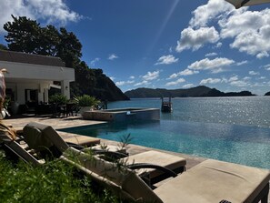A pool area with lounge chairs, a small boat, and a view of the sea and hills.