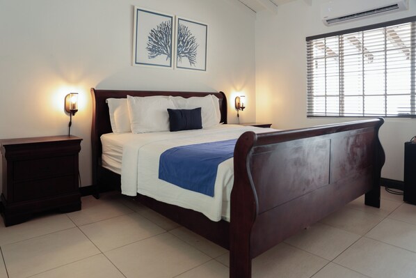 Cama bem arrumada com roupa de cama branca e azul, duas luminárias de cabeceira e uma janela com persianas.