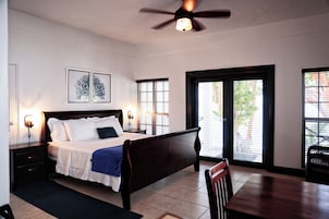 Quarto com cama, mesas de cabeceira, escrivaninha e cadeira. Há janelas com persianas e um ventilador de teto.