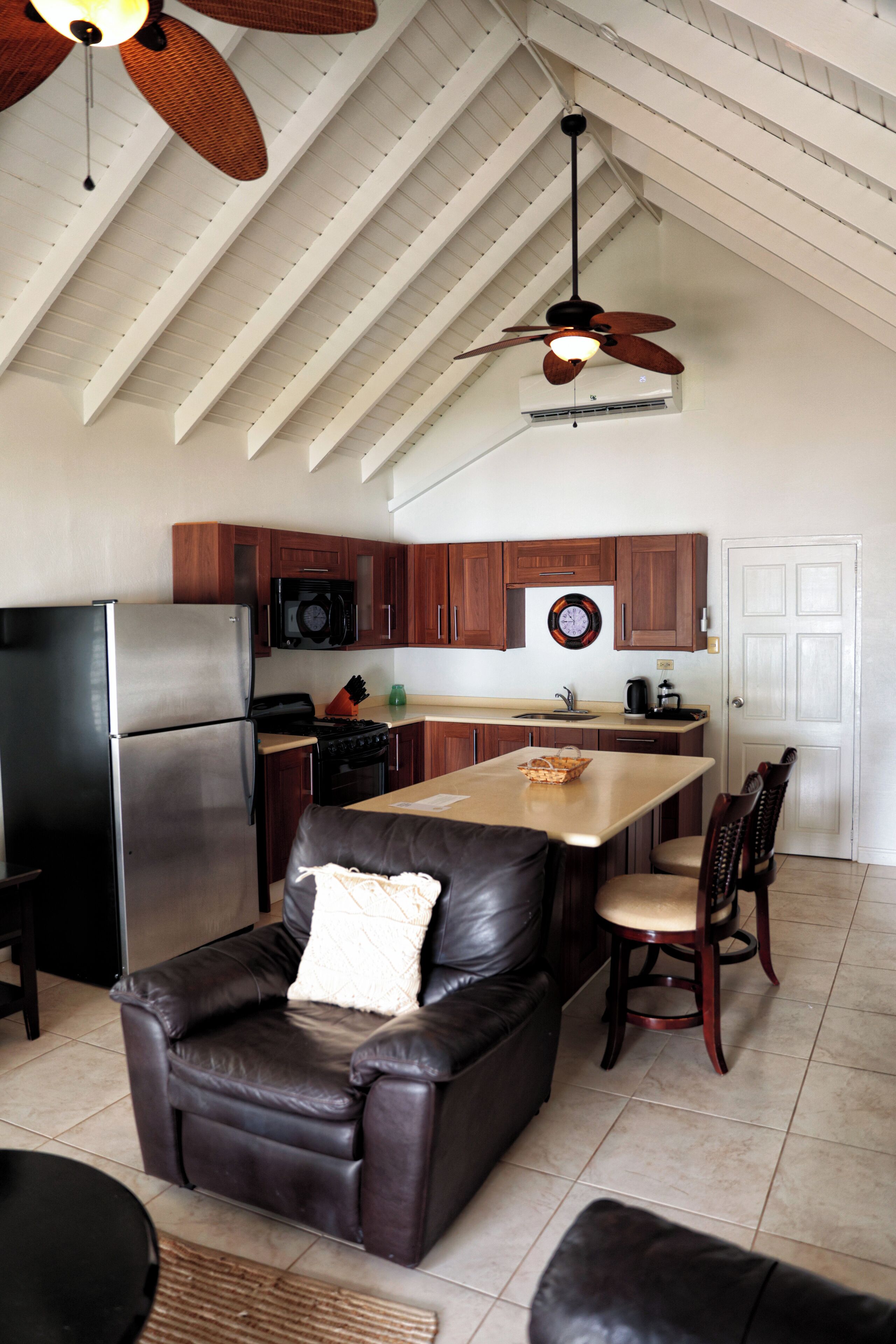 Una cocina con techo alto, armarios de madera, refrigerador, estufa, microondas y un comedor con sillas y mesa.