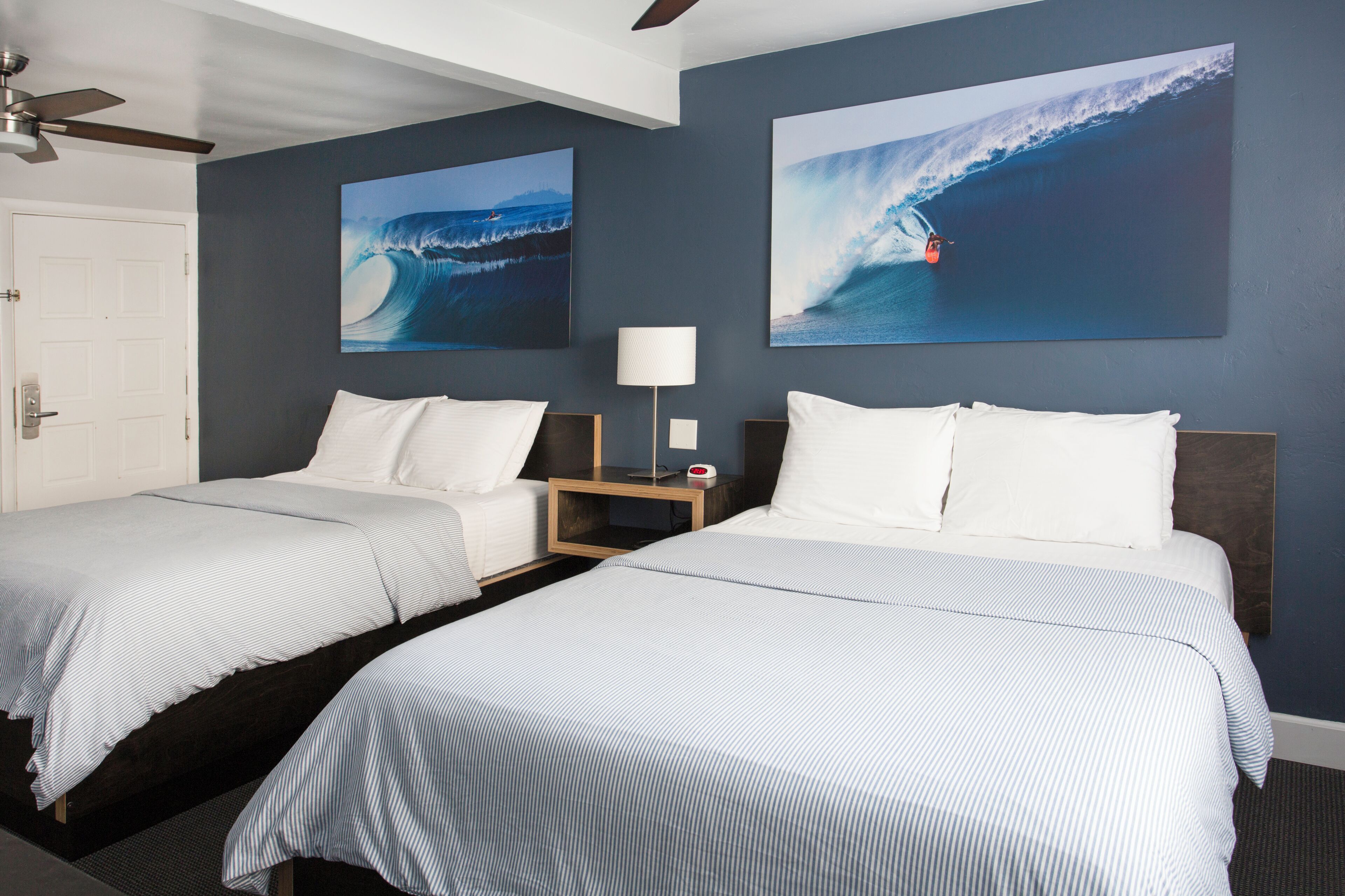 A hotel room with two beds, a ceiling fan, a nightstand with a lamp, and two large wave paintings on the wall.