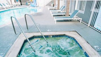 A hot tub with a metal ladder, lounge chairs, and a pool in the background.