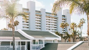 Una piscina con cerca blanca, un área de estar cubierta y un edificio con balcones y palmeras al fondo.