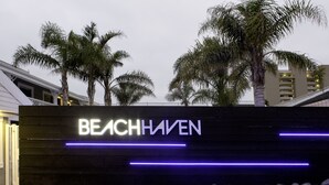 Un letrero con la inscripción iluminada 'BEACH HAVEN', rodeado de palmeras y un edificio moderno al fondo.
