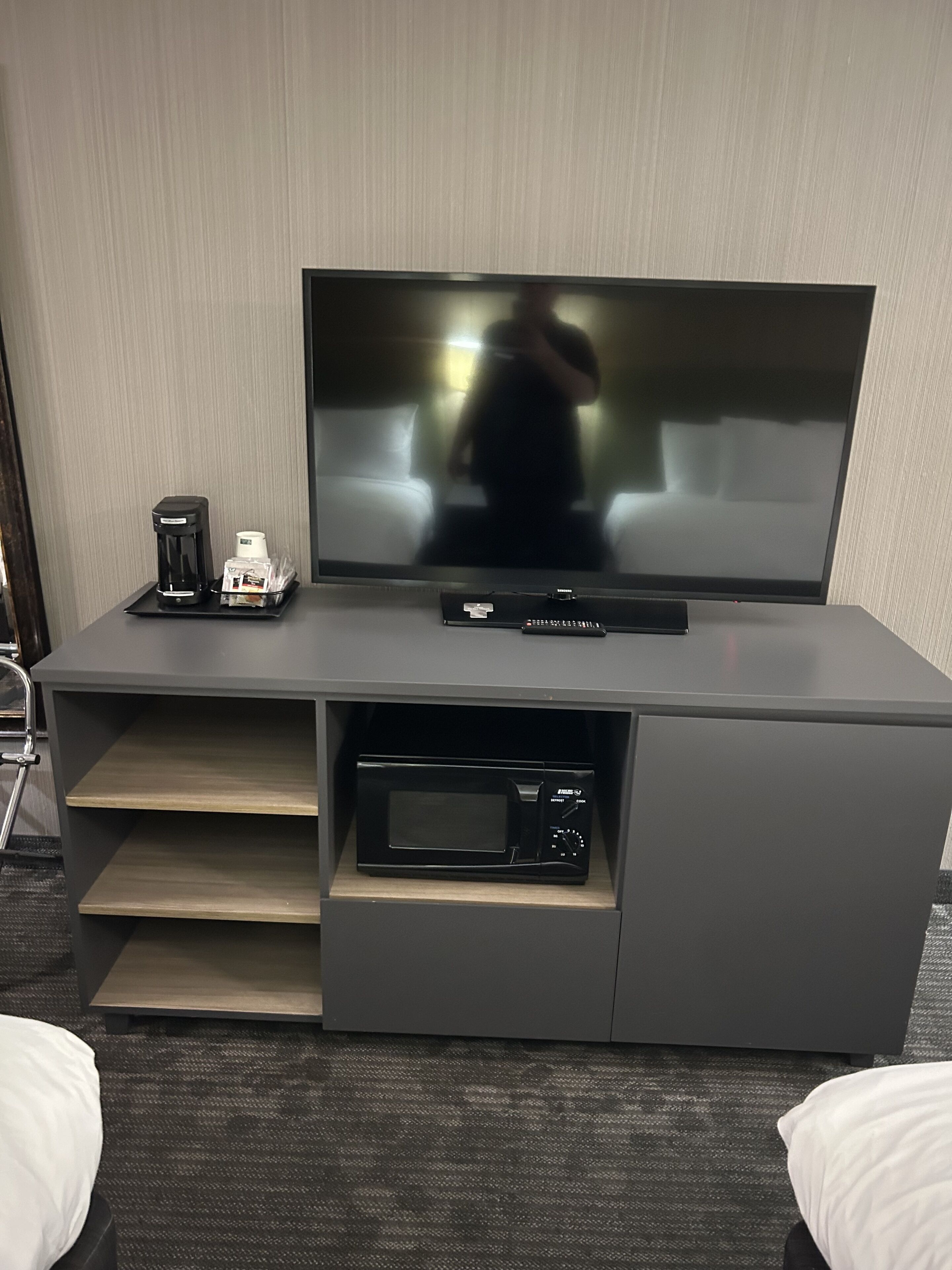 Ein modernes Hotelzimmer mit Flachbildfernseher, Kaffeemaschine und eingebauter Mikrowelle in einer grauen Unterhaltungsecke.