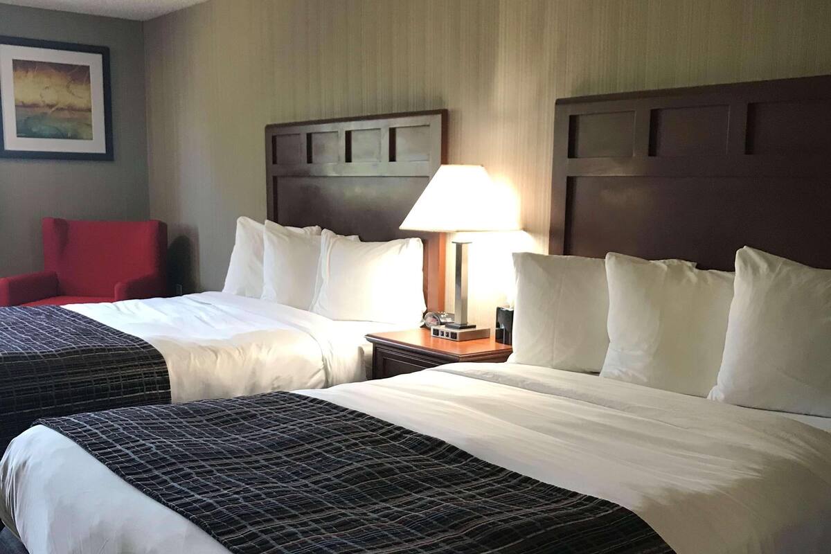 A hotel room with two beds, a red chair, a nightstand with a lamp, and a painting on the wall.