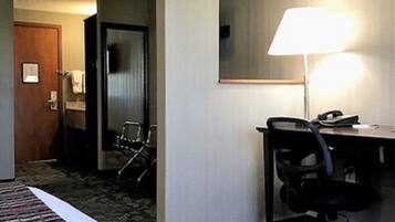 Quarto de hotel com cama, escrivaninha, cadeira, abajur e porta para um banheiro.