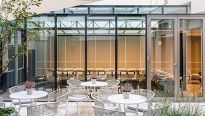 A modern outdoor seating area with round tables and wicker chairs, surrounded by glass walls and a metal structure. - Hotel Maximilian (Prague)