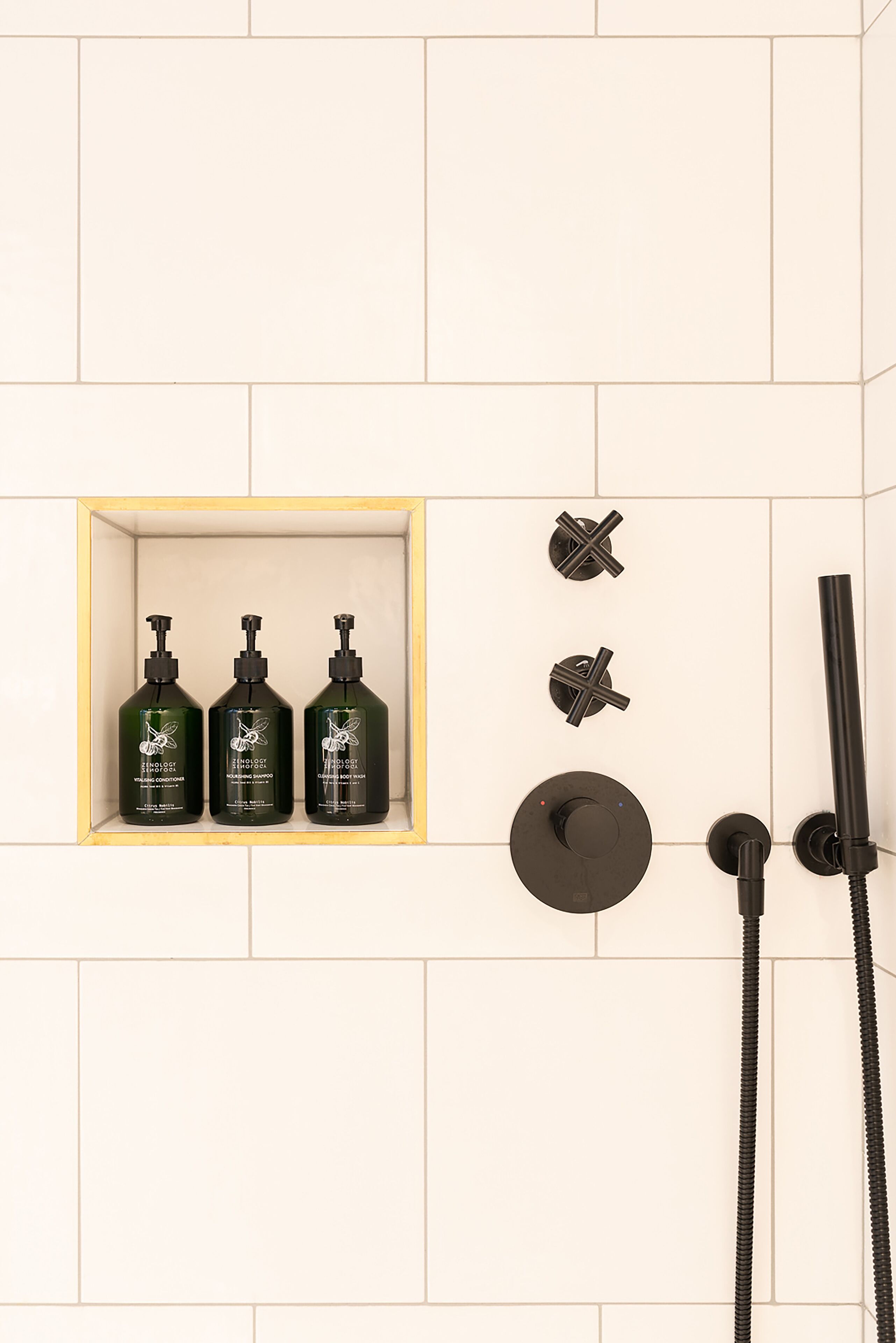 A shower area with three bottles of shampoo and conditioner, a handheld showerhead, and a wall-mounted control knob.
