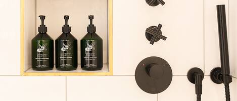 A shower area with three bottles of shampoo and conditioner, a handheld showerhead, and a wall-mounted control knob.